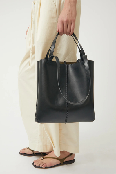 Cropped side-profile shot of a model carrying the black Brie Leon Reese Market Tote by its top handles, configured as a compact north-south tote with side press snaps fastened.