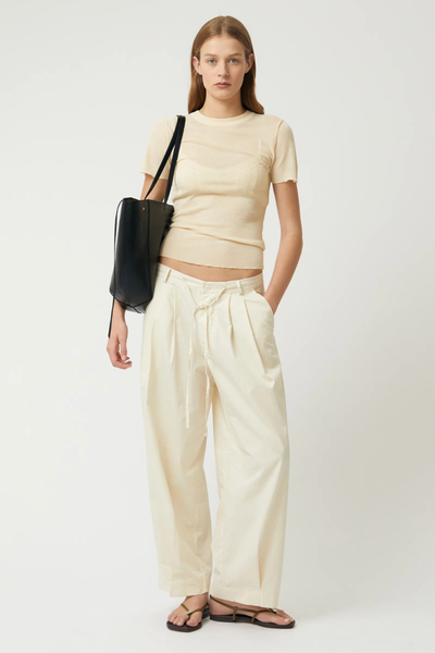Full-length studio shot of a model wearing a short-sleeve cream top, wide-leg cream trousers, and flat sandals, carrying the black Brie Leon Reese Market Tote as a compact shoulder bag.