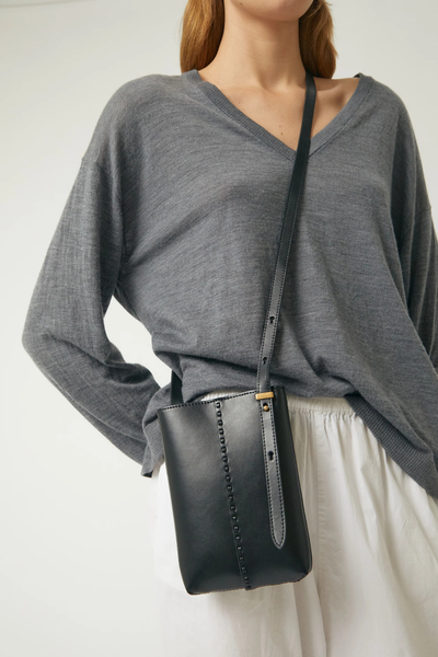 Cropped three-quarter portrait of a model wearing the Brie Leon Reese Pocket Bag in black recycled leather styled crossbody over a grey V-neck knit sweater and white trousers.