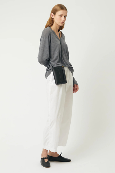 Full-length studio shot of a blonde model styled in a grey V-neck knit sweater, white wide-leg trousers, black ballet flats, and carrying the Brie Leon Reese Pocket Bag in black crossbody.