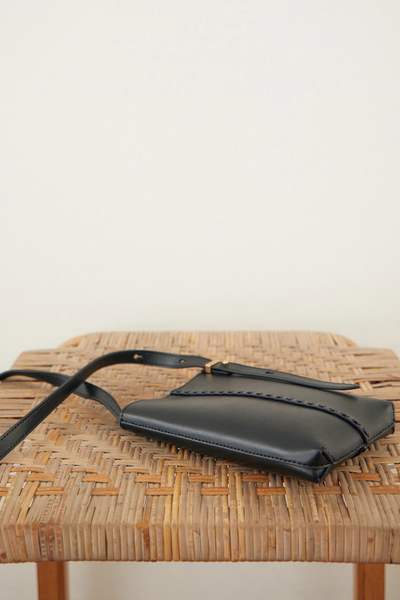 Lifestyle shot of the Brie Leon Reese Pocket Bag in black smooth recycled leather lying flat on a woven rattan stool against a plain light wall.