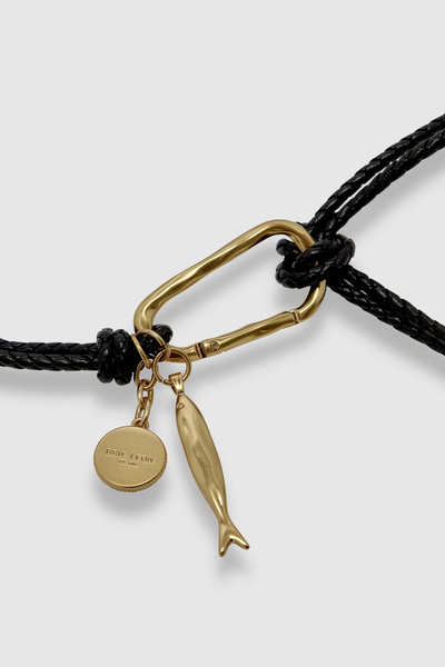 Close-up studio shot highlighting the organic-shaped gold carabiner, hand-braided black leather strap, and dangling fish and Brie Leon coin charms.