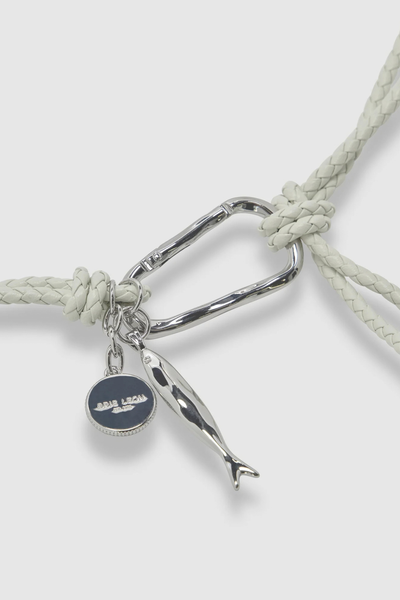 Close-up studio shot highlighting the organic-shaped silver carabiner, hand-braided elderflower-colored leather strap, and dangling fish and Brie Leon coin charms.