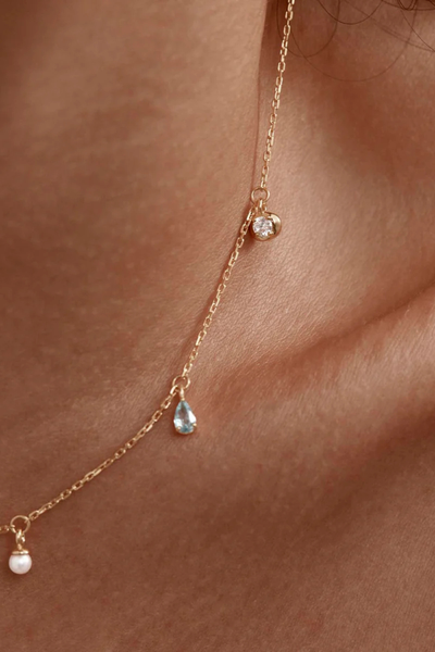 A close-up lifestyle campaign photograph of a model wearing the gold Breathe In Stillness Choker, capturing how the fine chain rests high on the neckline to exhibit the alternating drop charms—a round bezel-set cubic zirconia, a teardrop mint green nano gemstone, and a white freshwater pearl.