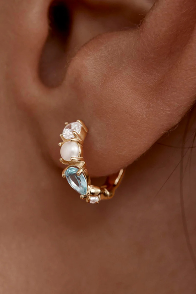 A macro close-up shot focusing on the gemstone alignment of a single gold vermeil hoop earring, detailing the polished prong settings holding a brilliant-cut clear cubic zirconia crystal, a small white freshwater pearl, and a vibrant teardrop-shaped light blue-green tourmaline gemstone.