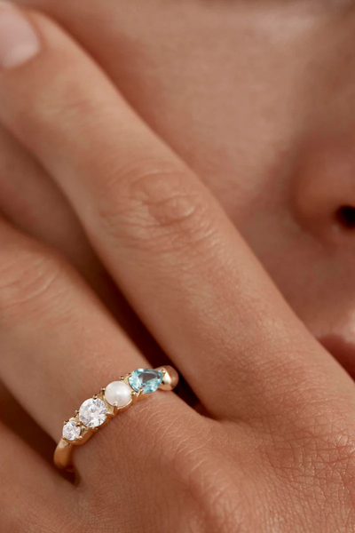 A lifestyle campaign photograph showing a close-up view of the gold Breathe In Stillness Ring being worn on a model's finger, highlighting how the multi-gemstone arrangement and its ocean-inspired colors sit gracefully against the hand.