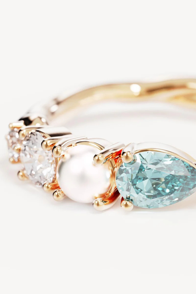 A macro close-up studio shot focusing on the stone settings of the gold vermeil statement ring against a white surface, detailing the polished gold prongs securing the faceted light blue-green tourmaline nano gemstone, the lustrous freshwater pearl, and the brilliant-cut cubic zirconias.