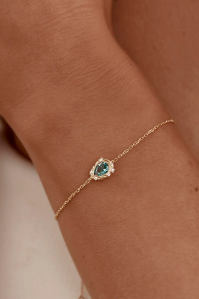 A minimal lifestyle campaign shot of the 18k gold vermeil fine chain bracelet worn on a model's wrist, showing the organic fluid shape and ocean-inspired blue-green hue of the gemstone pendant resting against the skin.
