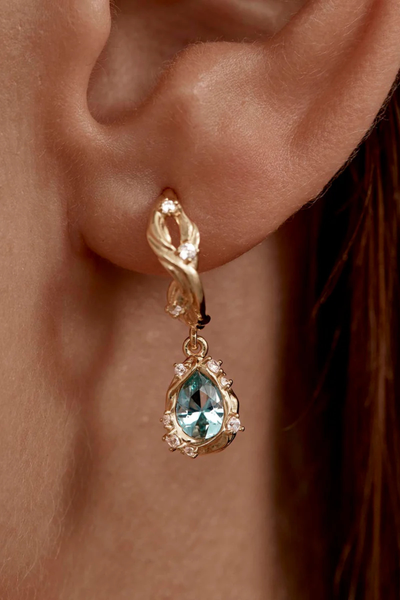 A macro lifestyle shot of the 18k gold plated hoop earring on a model's ear, focusing on the intricate details of the sparkling clear cubic zirconia crystals and the oceanic, magnetic blue-green hue of the central tourmaline nano gemstone pendant.