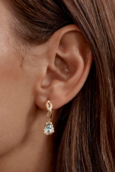 A lifestyle campaign image showing a close-up profile view of a model wearing the 18k gold plated Dive Deeper Hoop earring, highlighting its 10mm inner diameter and illustrating how the sea-green gemstone pendant catches the light when worn.