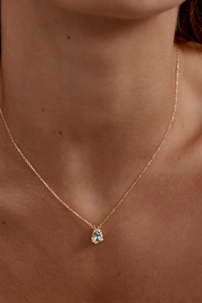 A macro, high-detail studio close-up of the 18k gold plated teardrop pendant resting on a seamless white surface, highlighting the shimmering facets of the central mint green tourmaline nano gemstone and the tiny cubic zirconias embedded along the undulating, wave-inspired metal border.