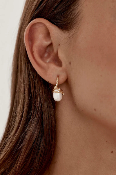 A soft-focus lifestyle image of the gold and baroque freshwater pearl drop earring on a model's ear, showcasing its scale, secure fit, and the delicate placement of tiny prong-set cubic zirconia crystals along the twisting gold vines.