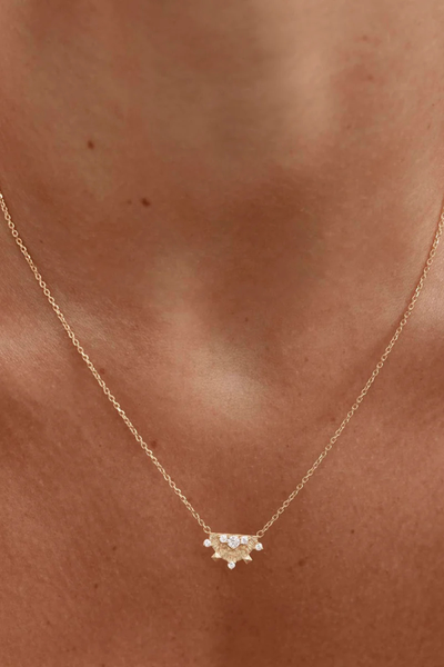 An ultra-close-up campaign image focusing on the fine craftsmanship of the gold vermeil Lotus Rain pendant against a warm, soft-focus brown backdrop, emphasizing the intricate radiating textures and the brilliant shimmer of the cubic zirconia settings.