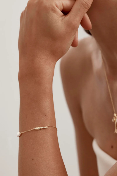 A soft-focus lifestyle campaign shot of the 18k gold vermeil Wishing Bracelet worn on a model's wrist, showing how the white freshwater pearl pendant and the polished gold anchovy charm drape elegantly against the skin, styled with a matching gold charm pendant necklace in the background.