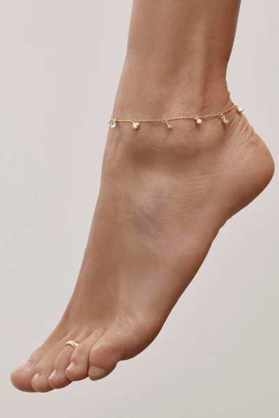 A high-angle lifestyle campaign shot featuring a model’s foot in profile against a neutral background, showcasing the 18k gold vermeil fine chain anklet resting gently on the ankle bone with dangling pearl and crystal charms, styled beautifully alongside a minimal gold band pinky toe ring.
