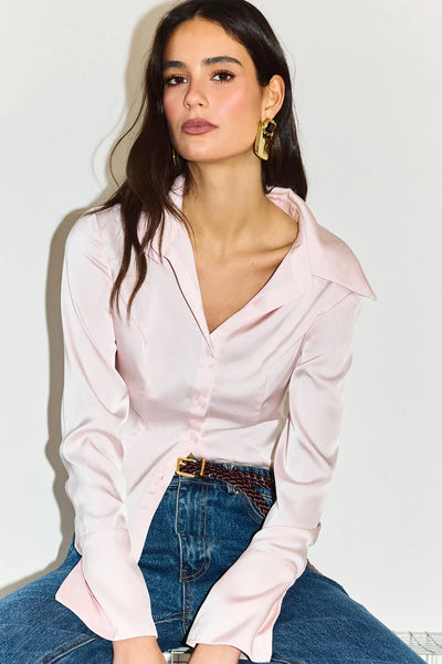 Close-up seated portrait of a model wearing the pink satin shirt tucked into blue denim jeans with a braided brown leather belt and gold statement earrings.