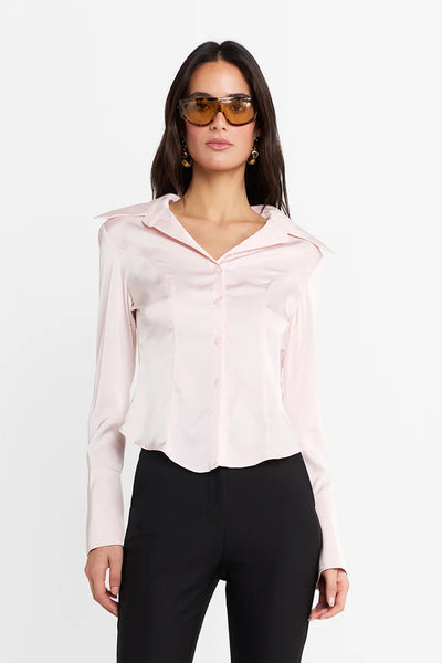 Waist-up studio view of a model wearing the pink fitted long-sleeve shirt, tinted amber sunglasses, statement earrings, and black pants, showcasing the structured collar and contoured princess seams.