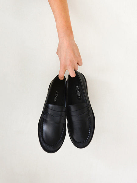Top-down view of a hand holding a pair of Senso Cooper III black leather loafers by the foot openings against a plain light background.