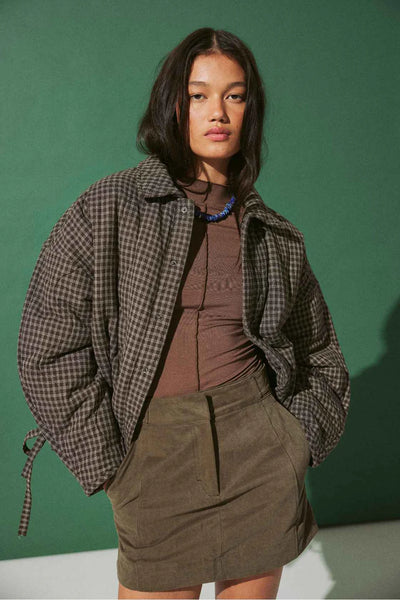 A studio portrait of a woman wearing the jacket open over a brown top and olive-green mini skirt against a dark green backdrop. A blue beaded necklace adds a pop of color.