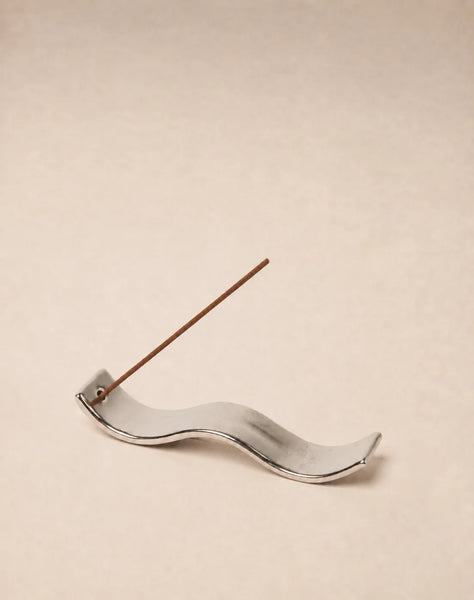 Studio product shot of the Gentle Habits Ceramic Wave Incense Holder featuring a metallic silver chrome glaze and a wave silhouette with an incense stick against a beige backdrop.