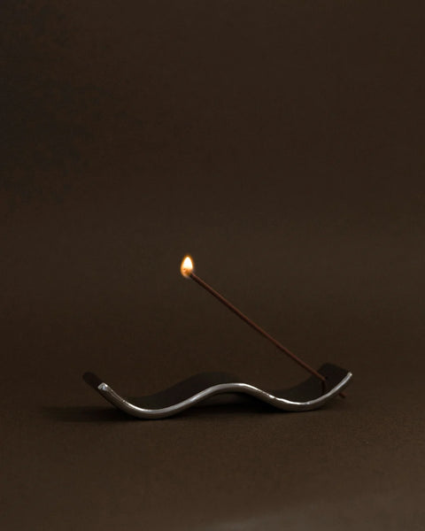 Side profile moody shot of the Gentle Habits Ceramic Wave Incense Holder in Silver holding a freshly lit burning incense stick against a dark background.