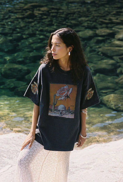 Outdoor location image of a model wearing the Kinga Csilla Desert Rose OG Tee in Black standing by a river, showing the full-length relaxed silhouette.