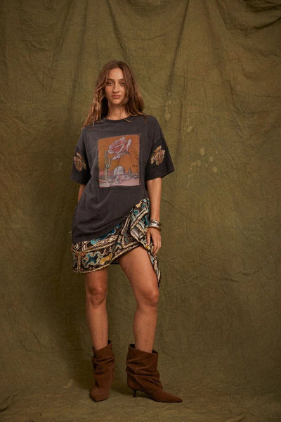 Studio full-length shot of a model styled in the oversized Kinga Csilla Desert Rose OG Tee in Black paired with a patterned mini sarong skirt and brown slouch boots.