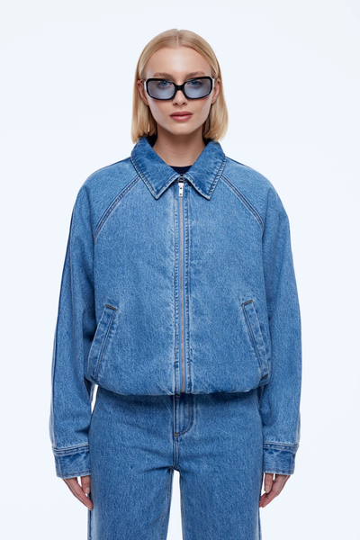 Three-quarter studio portrait of a model featuring the front zipped closure, elasticated hem, and front welt pockets of the ENA PELLY Billy Denim Bomber in Light Indigo.