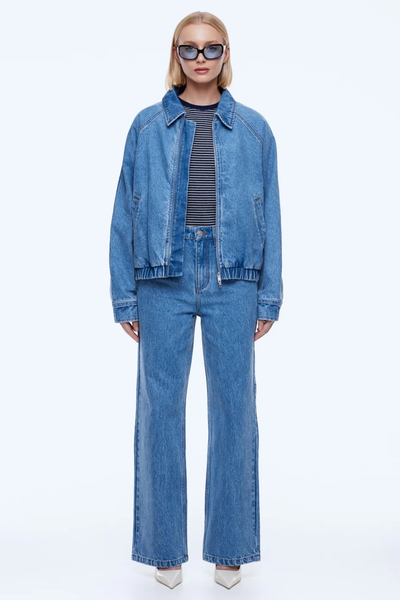 Full-body studio image of a model wearing the unzipped ENA PELLY Billy Denim Bomber in Light Indigo over a striped top and matching indigo jeans.