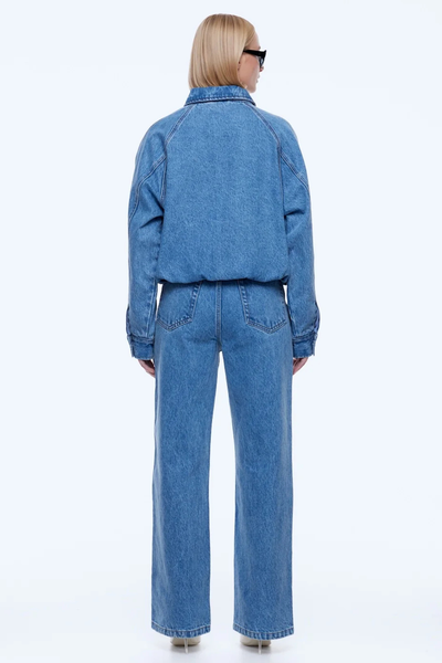 Rear full-length studio view of a blonde model showing the back silhouette, dropped raglan shoulder seams, and elastic waistband of the ENA PELLY Billy Denim Bomber in Light Indigo.