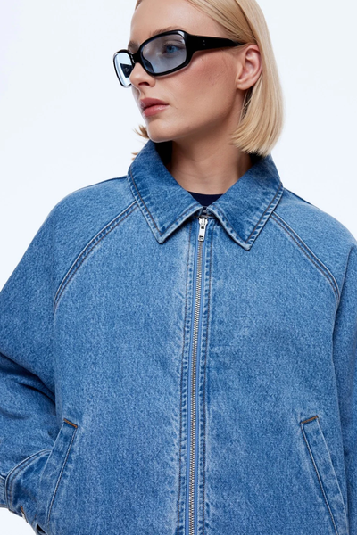 Close-up studio portrait of a blonde model wearing black sunglasses and the zipped-up ENA PELLY Billy Denim Bomber in Light Indigo showing raglan seam detailing and a classic collar.
