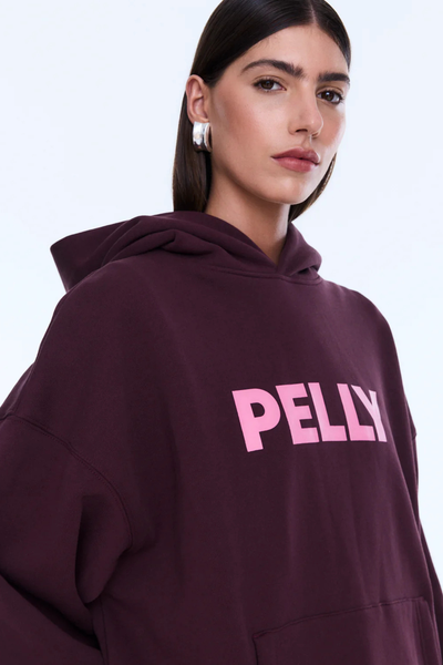 A full-length rear view of the model from behind, demonstrating the relaxed drape and clean backing of the deep cherry red fleece hoodie. The image highlights the structure of the self-lined hood resting flat against the shoulders and the ribbed hem, styled with dark-wash wide-leg denim jeans.