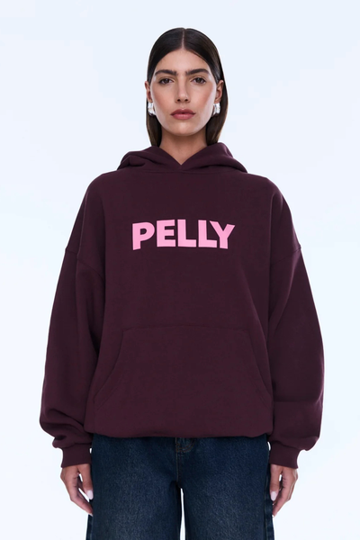 A full-length front view of the model standing straight while carrying a dark cherry red leather handbag. The photograph displays the complete oversized shape of the fleece hoodie, the contrast of the pink graphic, and the matching wide-leg dark denim jeans.