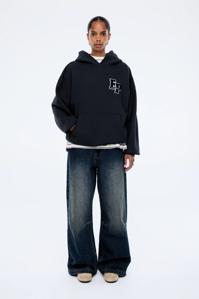 A full-length profile shot showing the side view of the model wearing the washed midnight hoodie. This perspective highlights the relaxed, slouchy shape of the fixed hood, the generous volume of the long sleeves, and the extended length of the dark-wash wide-leg denim jeans.
