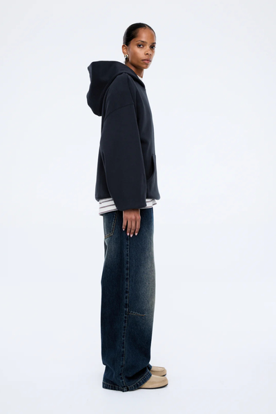 A full-length rear view of the model from behind, showcasing the clean, unadorned back paneling of the dark washed midnight hoodie. The shot illustrates the broad, oversized drop-shoulder fit and how the fixed hood sits flat across the upper back.