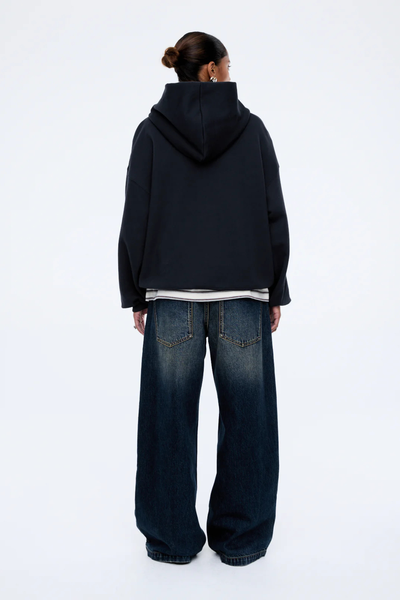 A full-length rear view of a model from behind, showcasing the clean back paneling of the Cassie Oversized Hoodie in washed midnight black. The photograph displays the broad drop-shoulder seam construction, the structural shape of the fixed hood resting flat against the upper back, and a peeking striped t-shirt hemline, styled with dark-wash wide-leg denim jeans.