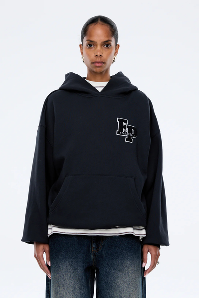 A cropped three-quarter front portrait focusing on the upper body of the model, detailing the soft texture of the dark cotton-blend fleece fabric, the comfortable structure of the fixed hood at the neckline, and the fine white-and-black embroidery lines of the collegiate chest graphic.