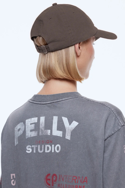 Rear view studio portrait of a model showing the back silhouette and adjustable metal buckle strap of the ENA PELLY Collegiate Cap in Washed Mocha.