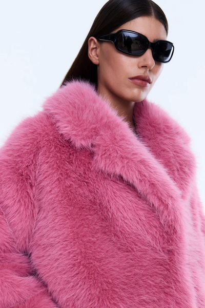 A full-length rear view of the model from behind, capturing the straight hemline and bold volume of the mid-length faux fur coat. The jacket is paired with dark-wash, wide-leg denim jeans.