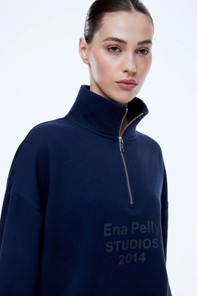 Close-up studio view of a model with hair slicked back wearing the ENA PELLY Kenzie 1/4 Zip Jumper in Navy, showcasing the silver quarter-zip high neck collar and tone-on-tone "Ena Pelly STUDIOS 2014" chest print.