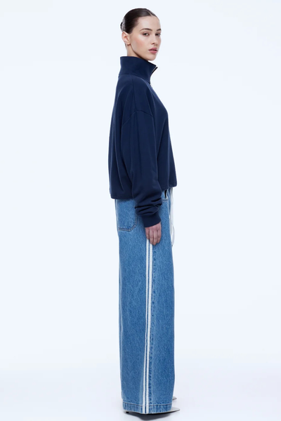 Side profile studio portrait of a model wearing the dark navy ENA PELLY Kenzie 1/4 Zip Jumper paired with wide-leg blue jeans with side stripes and pointed white heels.