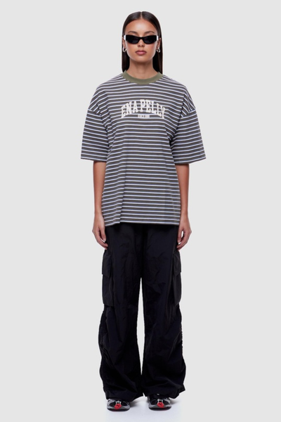 Full-length studio front shot of a model wearing dark sunglasses, an oversized green multi-striped graphic t-shirt, and relaxed black cargo pants.