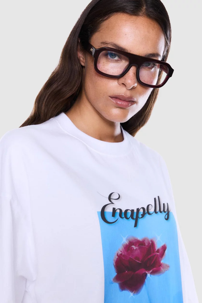 Close-up portrait of a model wearing black-framed glasses and a white crew-neck graphic tee featuring cursive branding above a pink rose graphic in a light blue rectangle.