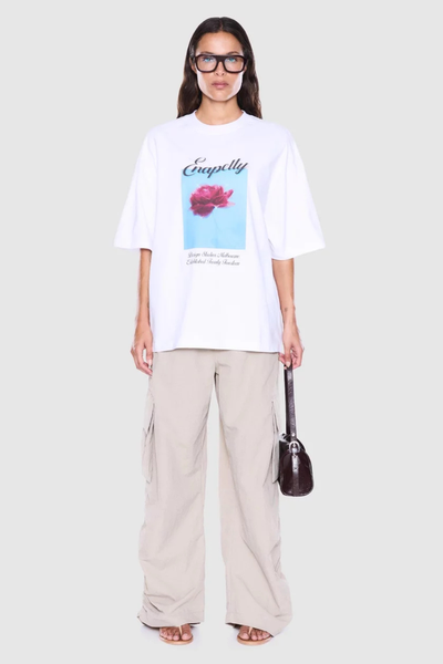 Full-length studio front shot of a model holding a dark mini shoulder bag, wearing an oversized white rose graphic tee and loose beige cargo pants.