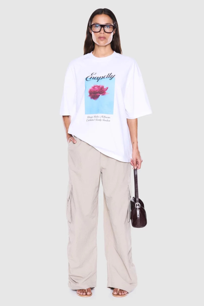 Full-length studio three-quarter front view of a model carrying a handbag, wearing a white oversized crew-neck graphic t-shirt paired with baggy beige trousers.