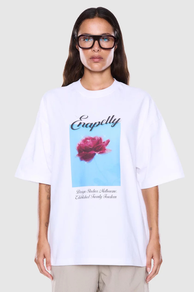 Studio front shot of a model wearing black-framed glasses and a white oversized graphic t-shirt featuring a blue box print with a pink rose and black cursive script text.