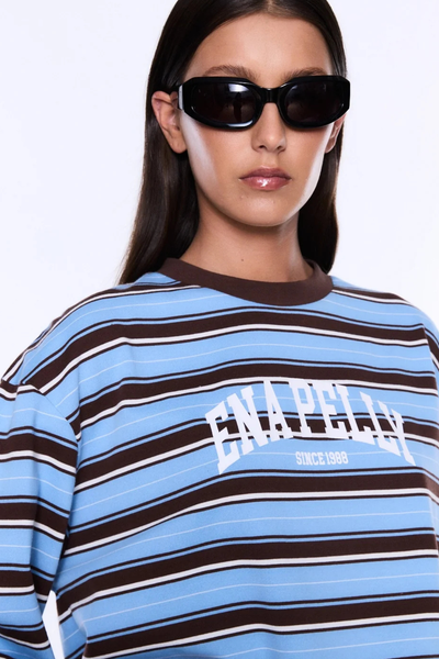 Close-up studio view of a brunette model wearing sunglasses and the ENA PELLY Maria Boxy Long Sleeve Tee showing the brown contrast crew neckline and white varsity logo print.