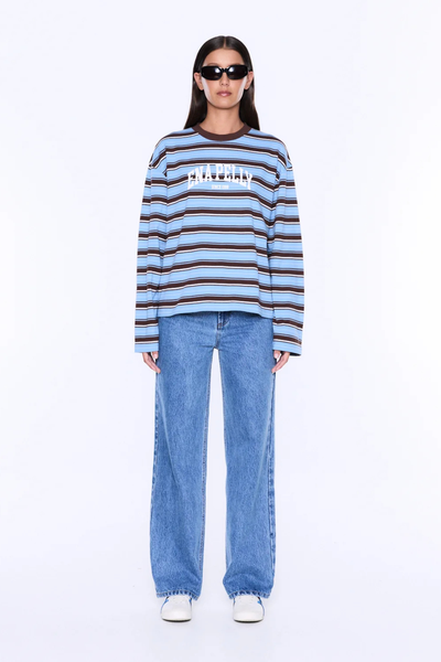 Full-length studio portrait of a model wearing the relaxed-fit ENA PELLY Maria Boxy Long Sleeve Tee in Blue/Brown Stripe with wide-leg blue jeans and white sneakers.