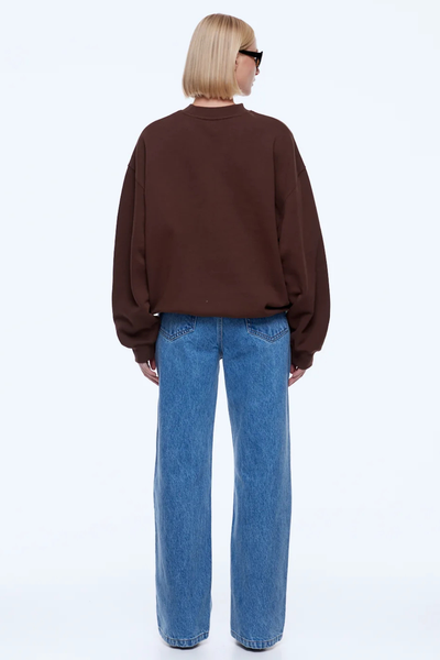 Full-length studio portrait of a model wearing the chocolate brown ENA PELLY Mila Oversized Sweater, styled with wide-leg blue jeans, sunglasses, pointed dark boots, and holding a dark handbag.