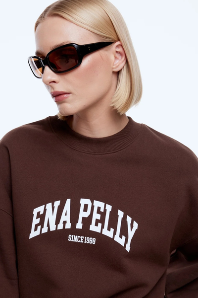 Close-up studio view of a blonde model wearing dark sunglasses and the chocolate brown ENA PELLY Mila Oversized Sweater, highlighting the white collegiate "ENA PELLY SINCE 1988" arched chest print and ribbed crew neckline.